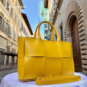 May include: A bright yellow tote bag with a woven design. The bag has two top handles and a detachable shoulder strap. The bag is sitting on a white surface in front of a European-style building.