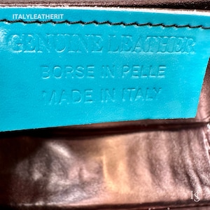 May include: A turquoise leather tag with the text "GENUINE LEATHER", "BORSE IN PELLE", and "MADE IN ITALY" in white lettering. The tag is attached to a brown leather surface.