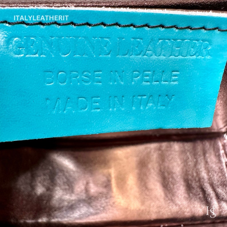 May include: A turquoise leather tag with the text "GENUINE LEATHER", "BORSE IN PELLE", and "MADE IN ITALY" in white lettering. The tag is attached to a brown leather surface.