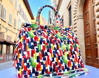 May include: A white leather handbag with a colorful abstract paint splatter design. The bag has a gold top handle and a detachable shoulder strap.