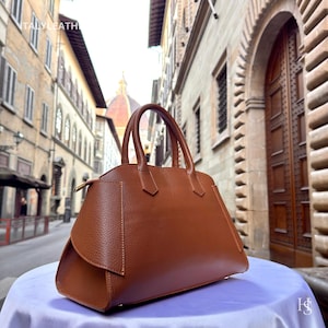 Puede incluir: Un bolso de mano de cuero marr&oacute;n con dos asas superiores. El bolso tiene una forma estructurada con un detalle de solapa en la parte delantera. El fondo muestra una escena de calle europea. El texto "ITALY LEATHER" es visible en la esquina superior izquierda.