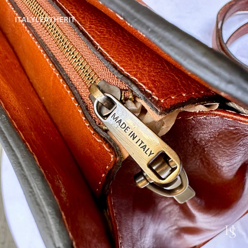 May include: Close-up of a brown leather bag with a zipper and a metal tag that reads "Made in Italy".
