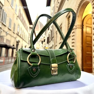 May include: A green leather handbag with two top handles and gold-colored hardware. The bag features a flap closure with a gold clasp and two large gold rings on the front. The bag is set against a blurred background of a European street.