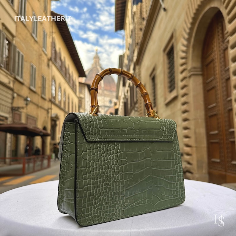 May include: A green crocodile embossed leather handbag with a bamboo handle. The bag is sitting on a white surface with a blurred background of an Italian city street.