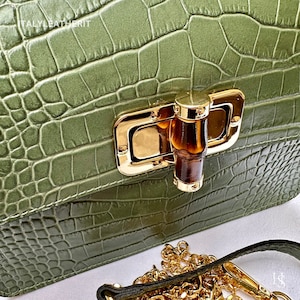 May include: A green crocodile embossed leather handbag with a gold and brown clasp. The bag has a gold chain strap.