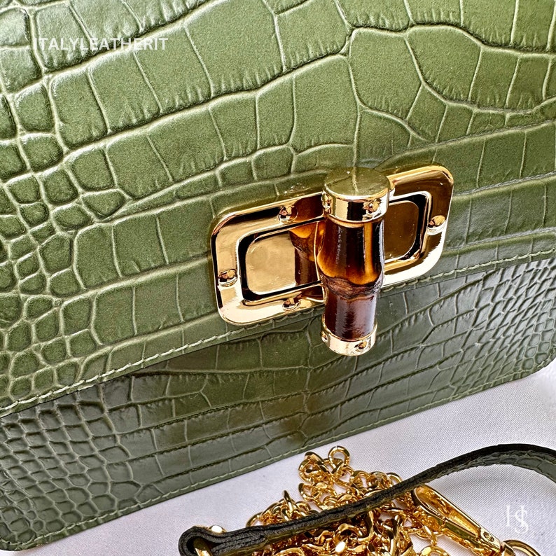 May include: A green crocodile embossed leather handbag with a gold and brown clasp. The bag has a gold chain strap.