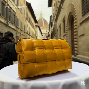 Puede incluir: Un bolso de mano de cuero amarillo mostaza con un dise&ntilde;o tejido. El bolso es rectangular con bordes redondeados y cierre de solapa. El bolso est&aacute; sobre una superficie blanca. El texto "ITALYLEATHERIT" es visible.