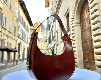 Könnte beinhalten: Eine halbmondförmige Handtasche aus braunem Leder mit Schulterriemen. Die Tasche hat einen Reißverschluss und Metallbeschläge. Der Hintergrund zeigt eine europäische Straßenszene mit Gebäuden und einer Kuppel.
