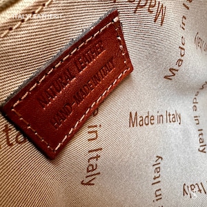 May include: Close-up of a brown leather tag with the text "NATURAL LEATHER HAND-MADE IN ITALY" stitched in white thread. The tag is on a beige fabric background with the text "Made in Italy" repeated in brown.