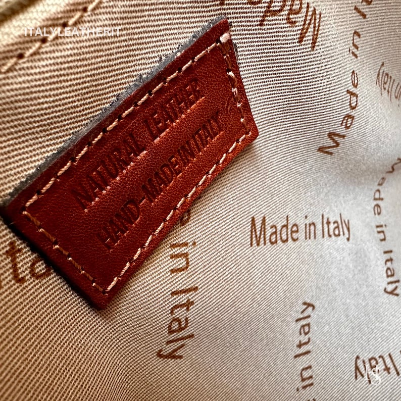 May include: Close-up of a brown leather tag with the text "NATURAL LEATHER HAND-MADE IN ITALY" stitched in white thread. The tag is on a beige fabric background with the text "Made in Italy" repeated in brown.