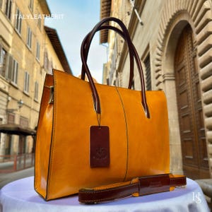 Peut inclure: Un sac fourre-tout en cuir jaune moutarde avec des poignées marron foncé et une bandoulière assortie. Le sac a une forme rectangulaire avec une étiquette attachée. Le sac est posé sur une surface blanche.