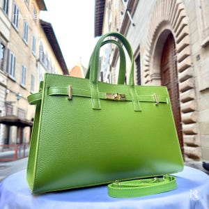 May include: A bright green leather handbag with a top handle and a detachable shoulder strap. The bag features gold-colored hardware, including a clasp closure and decorative accents. The bag is sitting on a white surface.