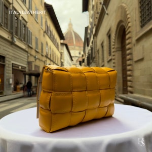 Puede incluir: Un bolso de mano de cuero amarillo mostaza con un dise&ntilde;o tejido. El bolso rectangular tiene un cierre de solapa y una correa de color neutro. El texto "ITALYLEATHERIT" es visible en la esquina superior izquierda.