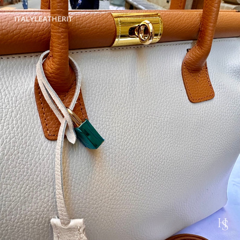 Puede incluir: Un bolso de mano de cuero blanco con ribete de cuero marr&oacute;n y una cerradura dorada. El bolso tiene una correa larga y un peque&ntilde;o candado sujeto al asa.