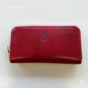 May include: A red leather wallet with a silver zipper and a small embossed emblem. The wallet is rectangular and has a smooth texture. The text "ITALYLEATHERIT" is visible at the top.