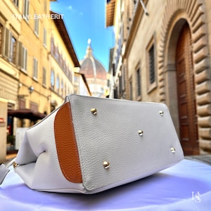 Puede incluir: Un bolso tote de cuero blanco con ribete de cuero marr&oacute;n. El bolso tiene detalles met&aacute;licos dorados y se muestra sobre una superficie blanca.