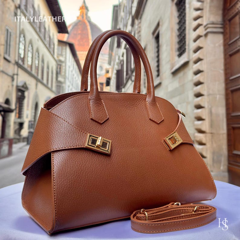 Puede incluir: Un bolso de mano de cuero marr&oacute;n con herrajes dorados, que incluyen un cierre de broche y detalles decorativos. El bolso tiene dos asas superiores y una correa para el hombro desmontable. El texto "ITALYLEATHER" es visible en la esquina superior izquierda.
