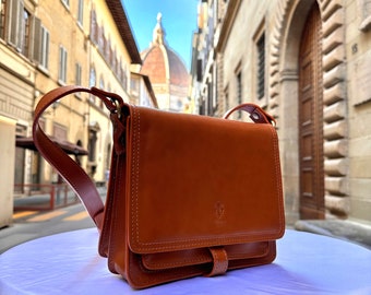 Può includere: Una borsa a tracolla in pelle marrone con chiusura a lembo e fibbia. La borsa ha un piccolo logo sul davanti ed è fotografata sullo sfondo di una città italiana.
