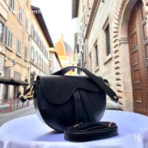 May include: A black leather saddlebag with a curved design and a detachable shoulder strap. The bag features gold-coloured hardware and is displayed on a white surface. The text "ITALY LEATHER" is visible in the upper left corner.
