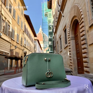 May include: A teal green leather handbag with a gold zipper and hardware. The bag has a detachable shoulder strap and a decorative gold ring detail. The bag is sitting on a purple surface, with a European city street in the background.