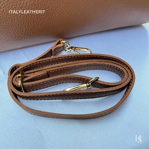 Puede incluir: Primer plano de una correa de hombro de cuero marr&oacute;n con cierres met&aacute;licos dorados. La correa est&aacute; enrollada frente a un bolso de cuero marr&oacute;n m&aacute;s grande. El texto "ITALYLEATHERIT" es visible en la esquina superior izquierda.