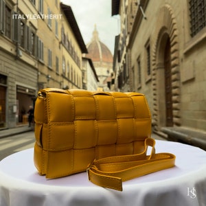 Puede incluir: Bolso bandolera de cuero tejido amarillo mostaza con una correa larga ajustable. El bolso es rectangular con un dise&ntilde;o tejido y un cierre dorado. El bolso est&aacute; sobre una superficie blanca con un fondo borroso de una calle europea.