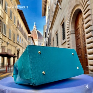 May include: Teal leather handbag with gold accents. The bag is shown on a blue surface with a blurry background of an Italian city.
