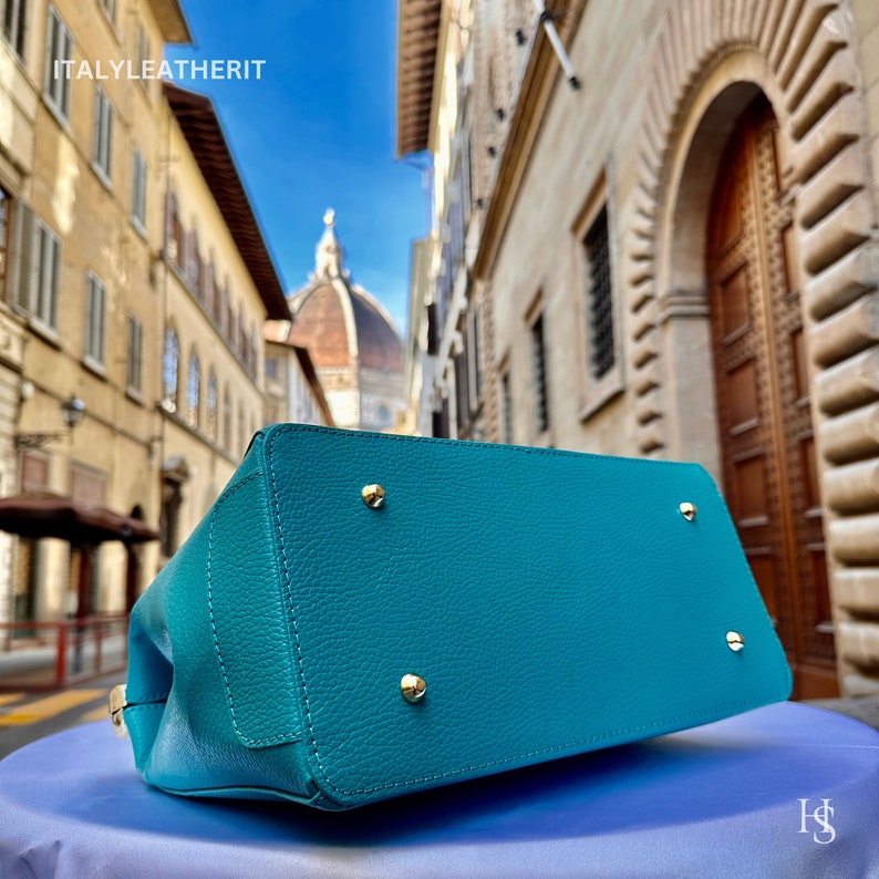 May include: Teal leather handbag with gold accents. The bag is shown on a blue surface with a blurry background of an Italian city.