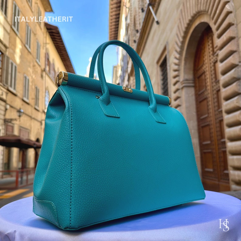 May include: A teal leather handbag with gold hardware. The bag has a top handle and a structured shape. The bag is photographed against a backdrop of a building in an Italian city.