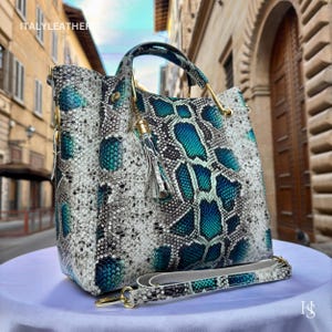 May include: A white and blue snakeskin handbag with a gold top handle and a tassel. The bag has a long strap and the text "ITALY LEATHER" is visible on the front.