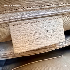 Puede incluir: Primer plano de una etiqueta de cuero blanco con el texto "GENUINE LEATHER" "BORSE IN PELLE" "MADE IN ITALY" impreso en ella.