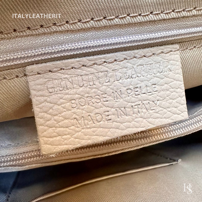 Puede incluir: Primer plano de una etiqueta de cuero blanco con el texto "GENUINE LEATHER" "BORSE IN PELLE" "MADE IN ITALY" impreso en ella.