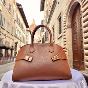 Puede incluir: Un bolso de mano de cuero marr&oacute;n con un dise&ntilde;o estructurado. El bolso presenta un asa superior, herrajes dorados y una forma rectangular. El bolso se exhibe sobre una superficie blanca con un fondo borroso de una calle de la ciudad.