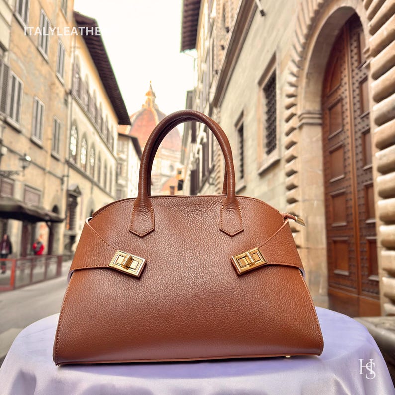 Puede incluir: Un bolso de mano de cuero marr&oacute;n con un dise&ntilde;o estructurado. El bolso presenta un asa superior, herrajes dorados y una forma rectangular. El bolso se exhibe sobre una superficie blanca con un fondo borroso de una calle de la ciudad.