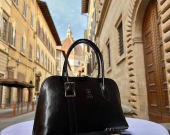 Könnte beinhalten: Eine schwarze Lederhandtasche mit einem oberen Griff und einem abnehmbaren Schulterriemen. Die Tasche hat eine rechteckige Form mit einer goldfarbenen Schnalle und Beschlägen. Die Tasche steht auf einer weißen Oberfläche mit verschwommenem Hintergrund einer europäischen Straße.