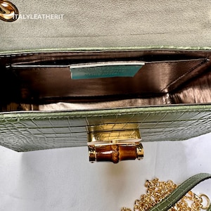 May include: A green crocodile embossed leather handbag with a gold bamboo-shaped clasp and a gold chain strap. The bag is open and the inside is lined with a brown fabric. The label inside reads "Genuine Leather, Made in Italy".