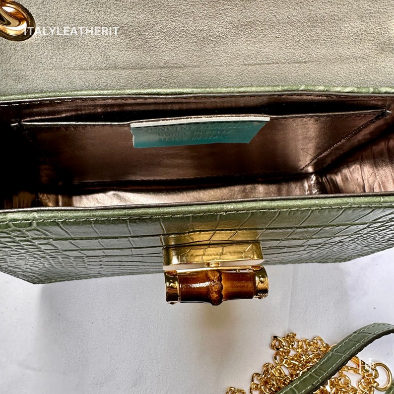May include: A green crocodile embossed leather handbag with a gold bamboo-shaped clasp and a gold chain strap. The bag is open and the inside is lined with a brown fabric. The label inside reads "Genuine Leather, Made in Italy".
