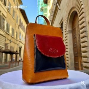 Puede incluir: Mochila de cuero en tonos marrón, rojo y azul marino. El cuerpo principal es marrón, con un bolsillo con solapa rojo y un bolsillo azul marino debajo. La bolsa tiene un asa superior y cierre de cremallera. El texto "ITALYLEATHERIT" es visible.