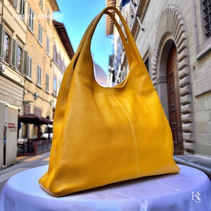 May include: A mustard yellow leather tote bag with long shoulder straps. The bag has a soft, slouchy design and a textured surface. The stitching is visible along the seams. The text "ITALY LEATHERIT" is visible at the top of the image.