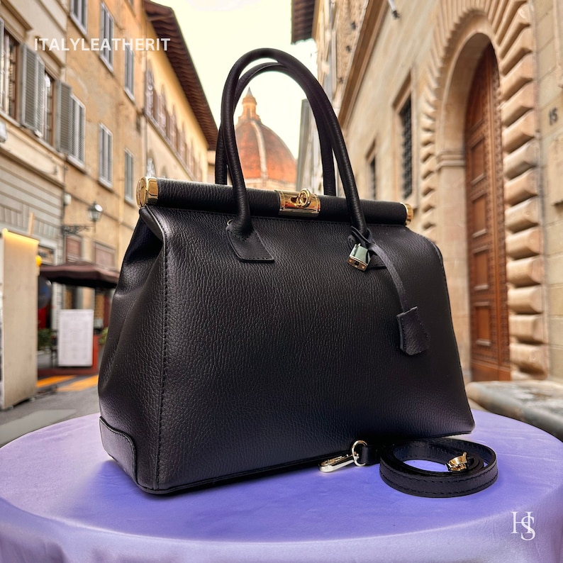 K&ouml;nnte beinhalten: Schwarze Ledertasche mit goldenen Beschl&auml;gen und einem abnehmbaren Schultergurt. Die Tasche hat einen oberen Griff und einen Schlie&szlig;verschluss. ITALYLEATHERIT steht oben auf der Tasche.