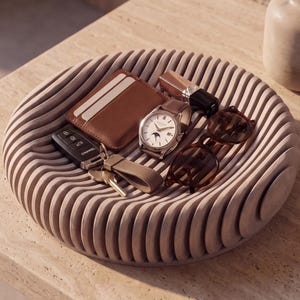 Peut inclure: Un plateau circulaire en bois beige clair avec une texture nervurée en forme de vagues. À l'intérieur du plateau se trouvent un porte-cartes en cuir marron, une montre-bracelet argentée à cadran blanc, une clé de voiture noire et des lunettes.