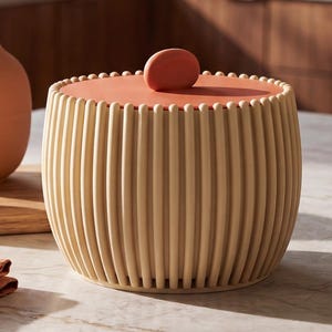 May include: A cylindrical storage container with a light beige slatted exterior and a flat, terracotta-coloured lid featuring a small, rounded handle. The container is positioned next to a terracotta vase, a small brass cup, and folded fabric napkins.
