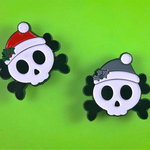 May include: Two enamel pins shaped like skulls with black crossbones behind them. One skull wears a red Santa hat with white trim and green holly leaves. The other skull wears a grey Santa hat with white trim and grey holly leaves.