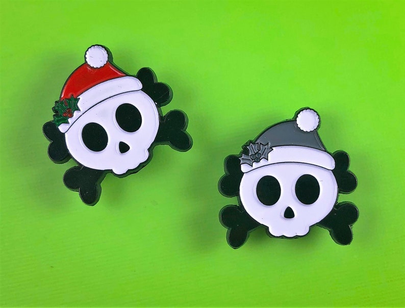 May include: Two enamel pins shaped like skulls with black crossbones behind them. One skull wears a red Santa hat with white trim and green holly leaves. The other skull wears a grey Santa hat with white trim and grey holly leaves.
