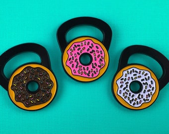 May include: Three enamel pins shaped like black kettlebells with donut centers. One features a brown donut with multicolored sprinkles, one a pink donut with green sprinkles, and one a white donut with multicolored sprinkles on a yellow base.
