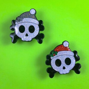 May include: Two skull-shaped enamel pins with black crossbones. One skull wears a dark gray glitter Santa hat with holly, and the other wears a red glitter Santa hat with green holly leaves and red berries. Both skulls have a white glitter finish.