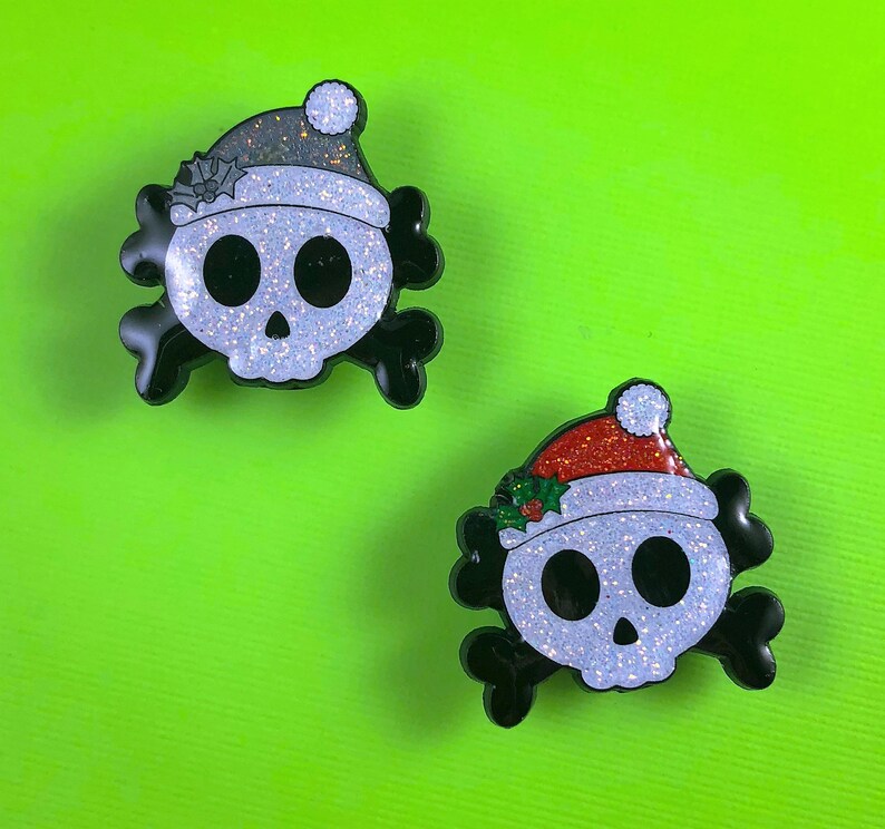 May include: Two skull-shaped enamel pins with black crossbones. One skull wears a dark gray glitter Santa hat with holly, and the other wears a red glitter Santa hat with green holly leaves and red berries. Both skulls have a white glitter finish.