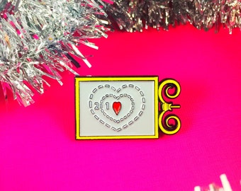 May include: A rectangular enamel pin with a yellow border and a light gray center. The design features two concentric dashed heart outlines, the numbers 2 and 1, and a small red heart. A yellow decorative scroll element is attached to the right side.