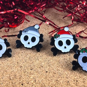 May include: Four enamel pins shaped like skulls with crossbones. Two skulls wear glittery silver Santa hats, one wears a solid white hat, and one wears a red glittery Santa hat. Each hat features a small green holly leaf accent with red berries.