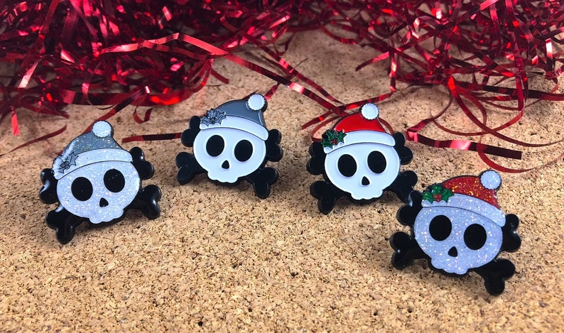 May include: Four enamel pins shaped like skulls with crossbones. Two skulls wear glittery silver Santa hats, one wears a solid white hat, and one wears a red glittery Santa hat. Each hat features a small green holly leaf accent with red berries.